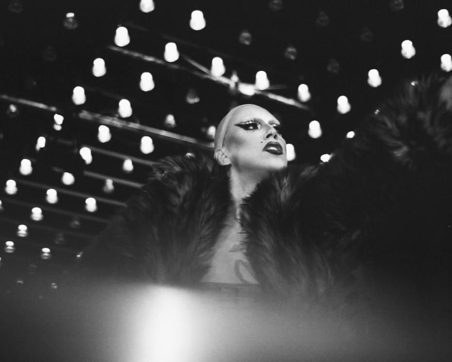 Artiste drag en fourrure, sous un plafond d’ampoules.